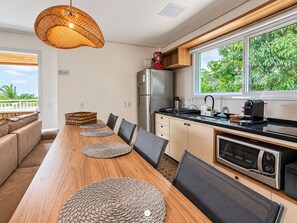 A modern kitchen with a long wooden table, black chairs, a built-in oven, and a large window with a view of greenery. - Villa Verde Bangalô - Garopaba - SC (Garopaba)
