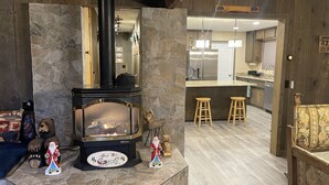 A cozy living room with a stone fireplace, wooden accents, and a kitchen area visible through an open doorway.