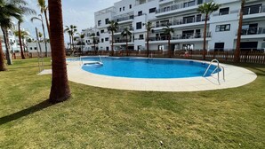 Un moderno edificio de apartamentos con piscina rodeada de palmeras y un área verde.