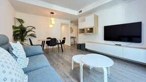 Una sala moderna con un sofá gris, dos mesitas de noche blancas, un área de comedor con una mesa y sillas, un televisor montado en la pared y una planta en maceta.