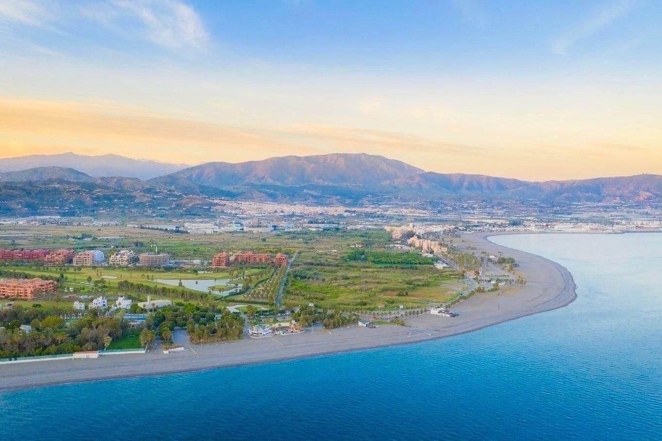 An aerial view of a coastal city with a beach, residential buildings, and mountains in the background.