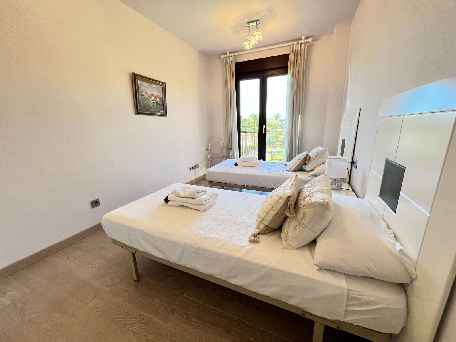A bedroom with a large bed, a smaller bed, a window with curtains, a ceiling light, and a framed picture on the wall.