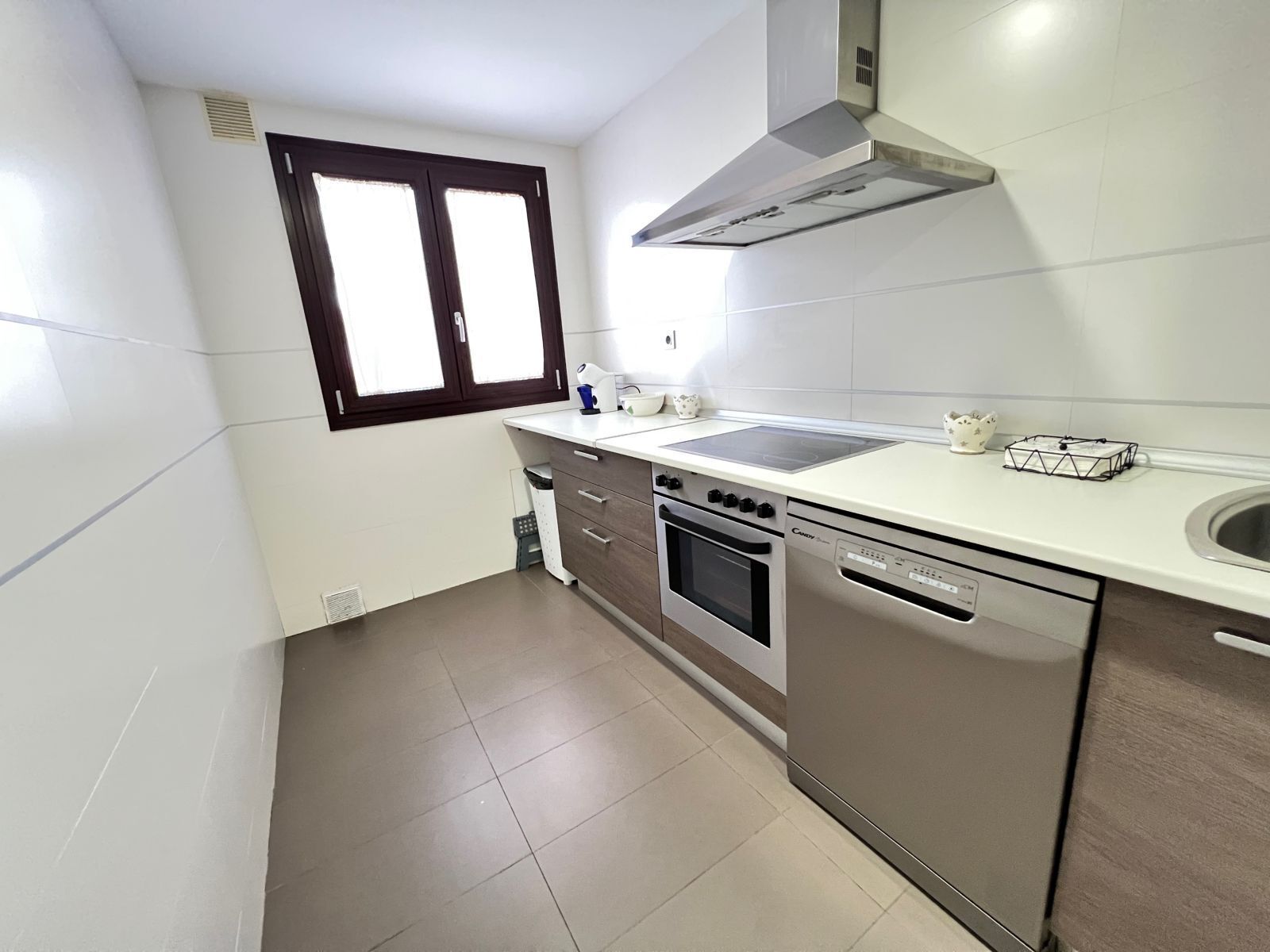 Cocina compacta con estufa, horno y lavavajillas, una ventana con persianas y un salpicadero blanco de azulejos.