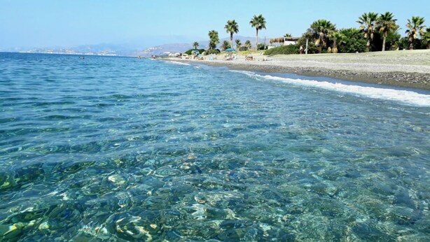 Une plage à l’eau cristalline, bordée de palmiers, avec une chaîne de montagnes qui se dessine à l’horizon.