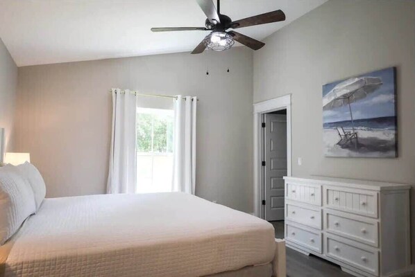 Un dormitorio con cama, ventilador de techo, ventana con cortinas, cómoda y un cuadro en la pared.