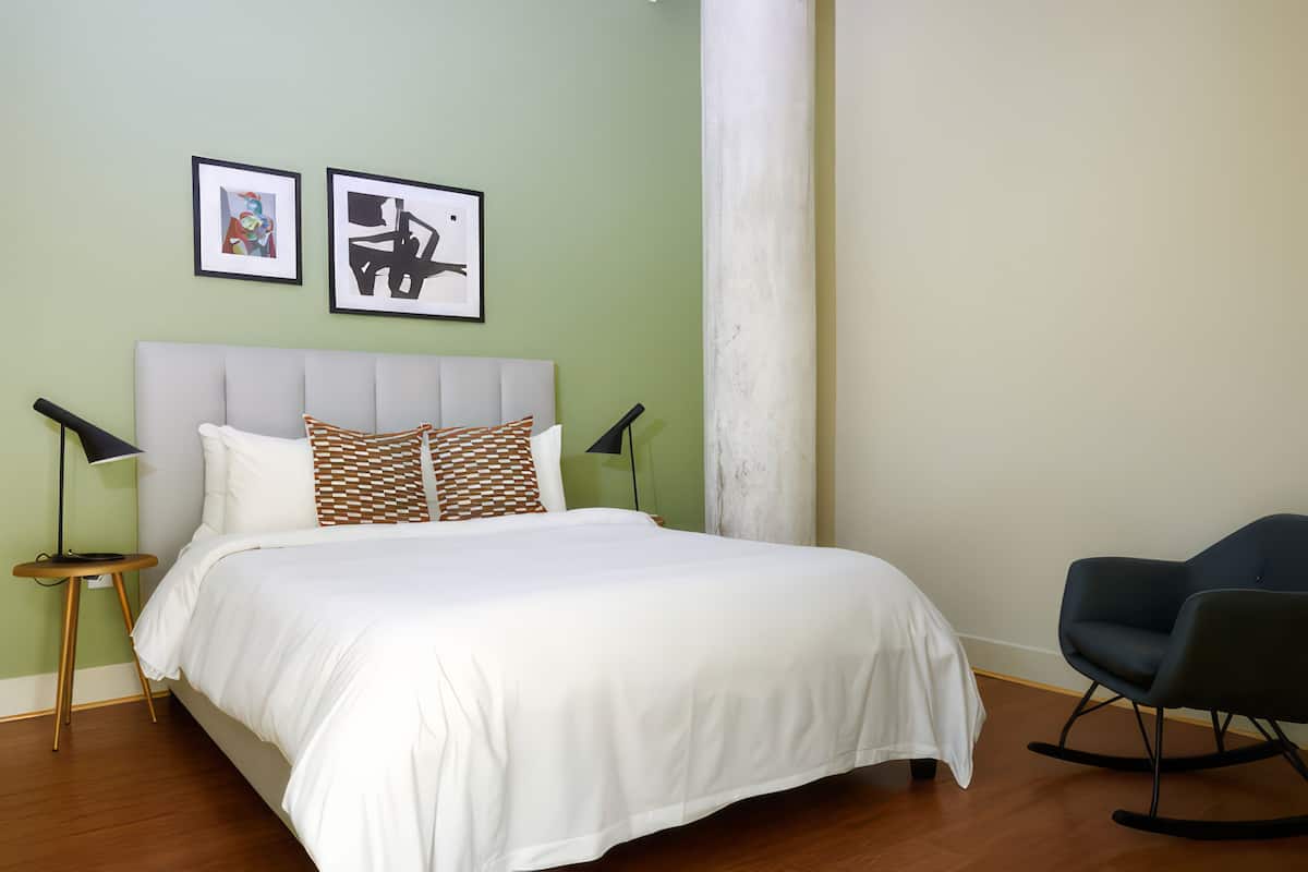 Un lit bien fait, avec du linge de lit blanc et des coussins à motifs, une chaise moderne et deux œuvres d’art encadrées accrochées au mur.