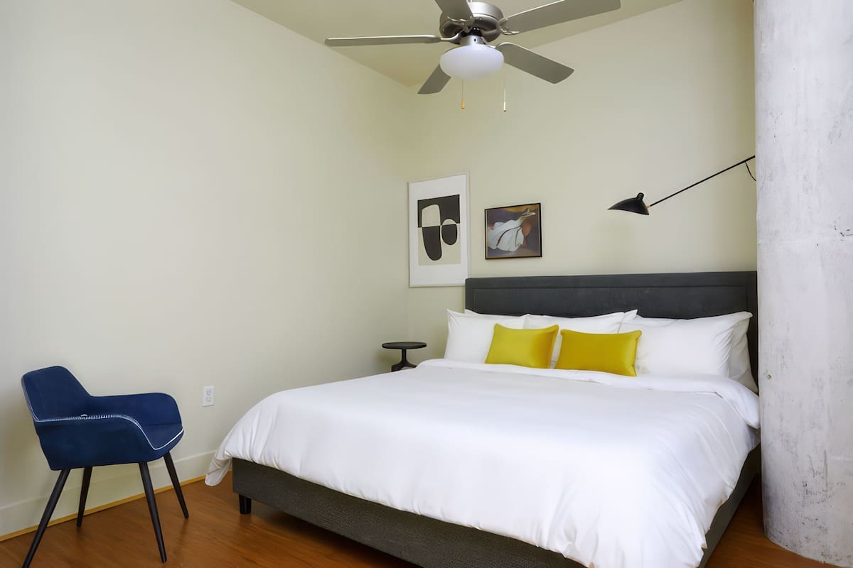 Une chambre à coucher comprenant un lit, une chaise, un ventilateur de plafond, une table de chevet et des œuvres d’art accrochées au mur.