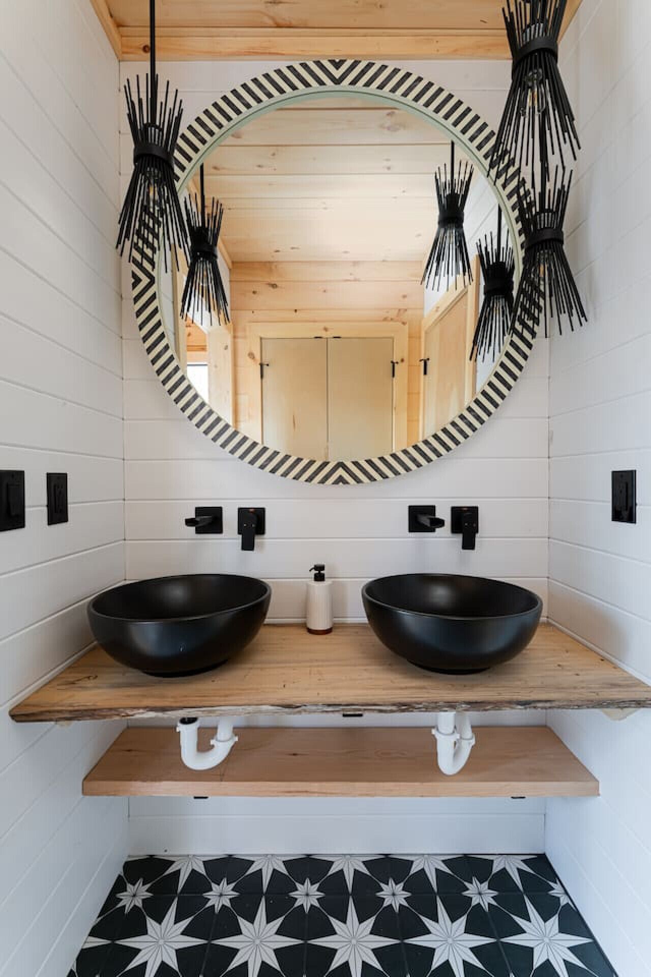 A bathroom with two black sinks, a wooden shelf, a mirror with a black and white frame, and a patterned floor.