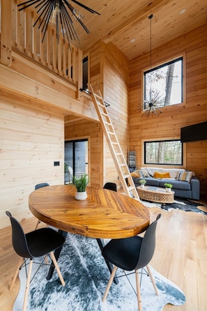 Un interior de madera con una mesa redonda de madera, sillas negras, un sofá y una escalera que lleva a una zona de entrepiso.