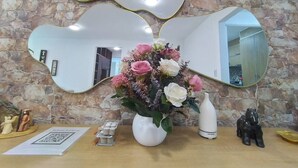 En spegel med en vågig ram, en vas med rosa och vita blommor och en liten statyett på ett träbord mot en stenmur.