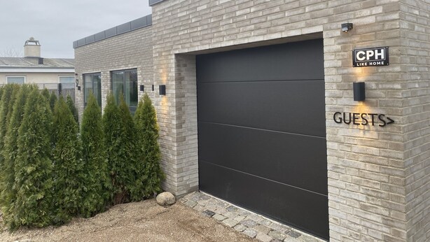 Un edificio moderno con fachada de ladrillo, una puerta de garaje oscura y un letrero que indica 'CPH Like Home' y 'GUESTS'.