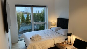 Un dormitorio con cama, escritorio con computadora portátil, lámpara y vista a árboles y un edificio a través de un ventanal grande.