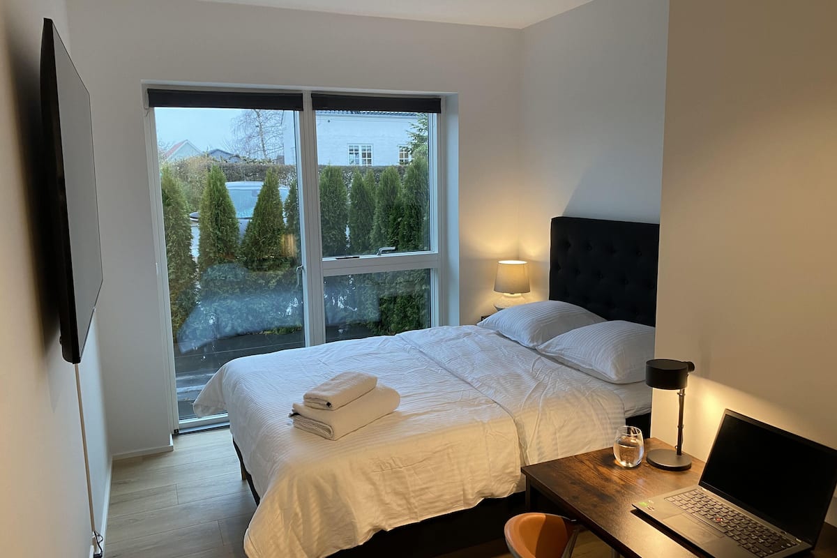A bedroom with a bed, a desk with a laptop, a lamp, and a view of trees and a building through a large window.