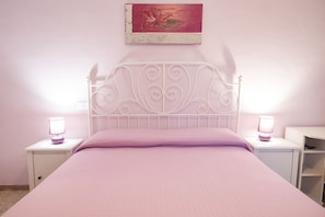 Una cama con cabecera de metal blanco, dos mesitas de noche con lámparas, una colcha rosa y un cuadro en la pared.