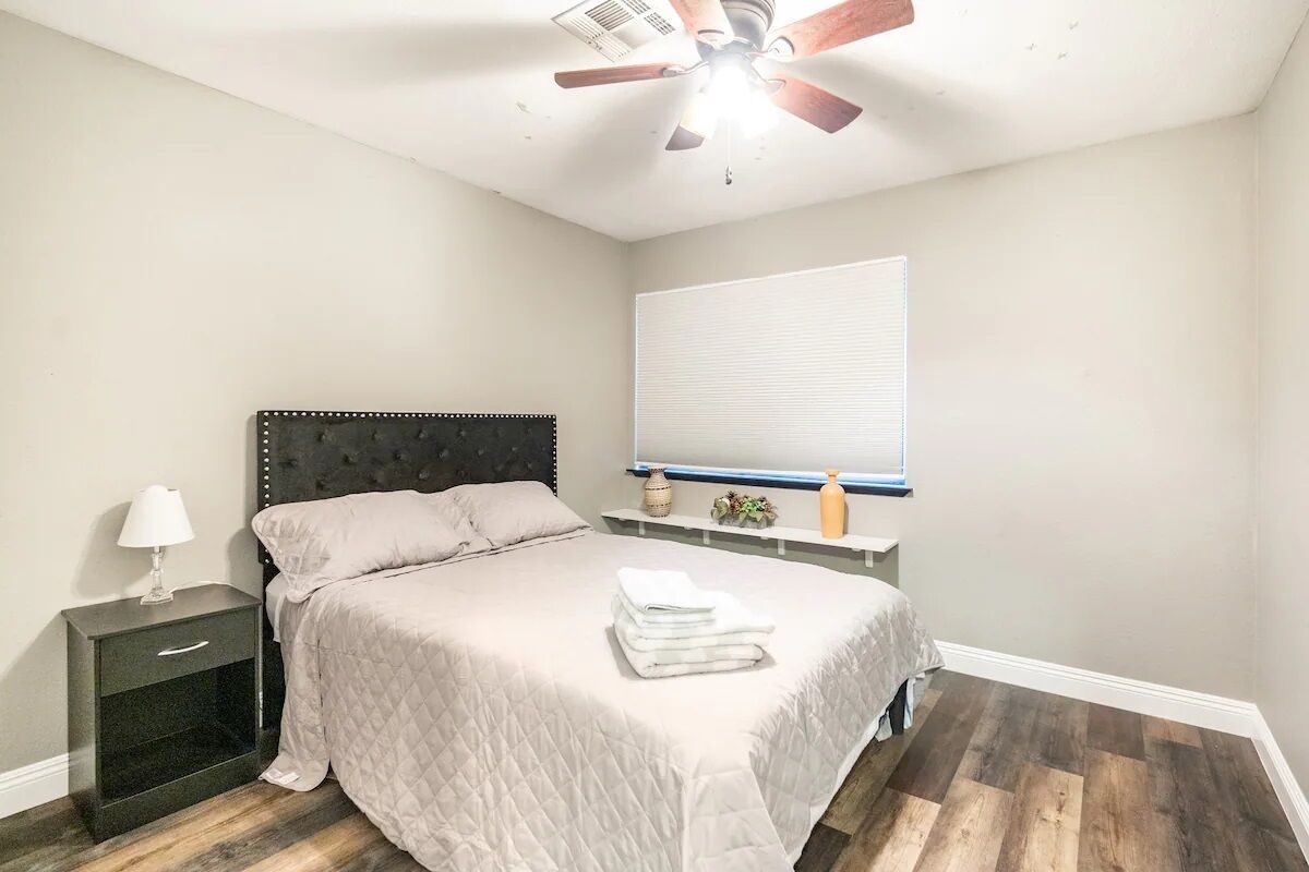 Un dormitorio con cama, mesita de noche, lámpara, ventilador de techo, ventana y una planta pequeña.