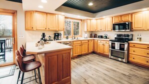 A modern kitchen with wooden cabinets, a central island with stools, stainless steel appliances, and a large window providing natural light. - FallWater - 5BR Lakehouse, WtrFrnt, Ht Tub, F/pit. Close to Ski and Golf! (Boyne City)