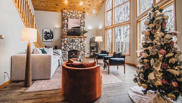 A cozy living room with a stone fireplace, a Christmas tree, and a large window. - FallWater - 5BR Lakehouse, WtrFrnt, Ht Tub, F/pit. Close to Ski and Golf! (Boyne City)