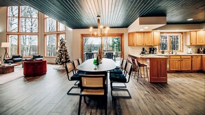 A spacious dining area with a wooden table and chairs, a Christmas tree, and large windows offering a view of the outdoors. - FallWater - 5BR Lakehouse, WtrFrnt, Ht Tub, F/pit. Close to Ski and Golf! (Boyne City)