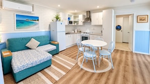 A compact living space with a kitchenette, a sofa, a dining table with chairs, and a view of a beach through a window.