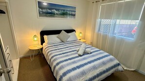 Een net opgemaakt bed met een gestreept deken, twee bedlampen, een ingelijst beeld van golven en een raam met gordijnen.
