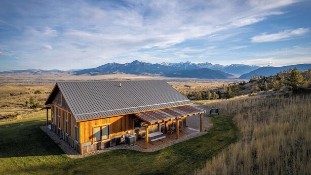 Una cabaña de madera con techo metálico, rodeada de césped y con vistas a montañas al fondo.