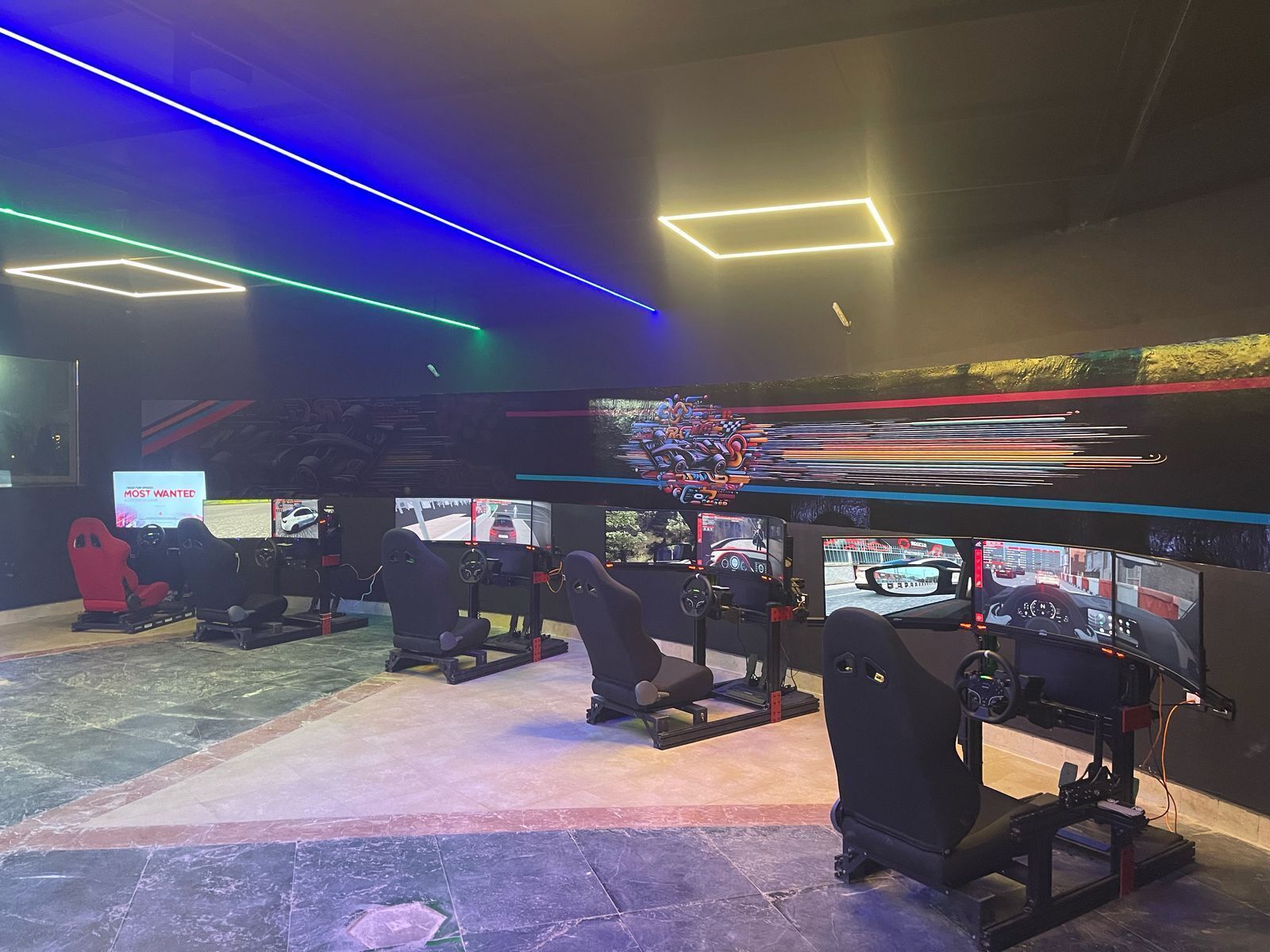 A gaming area with multiple racing simulators, a large screen displaying a racing game, and neon lighting on the ceiling.