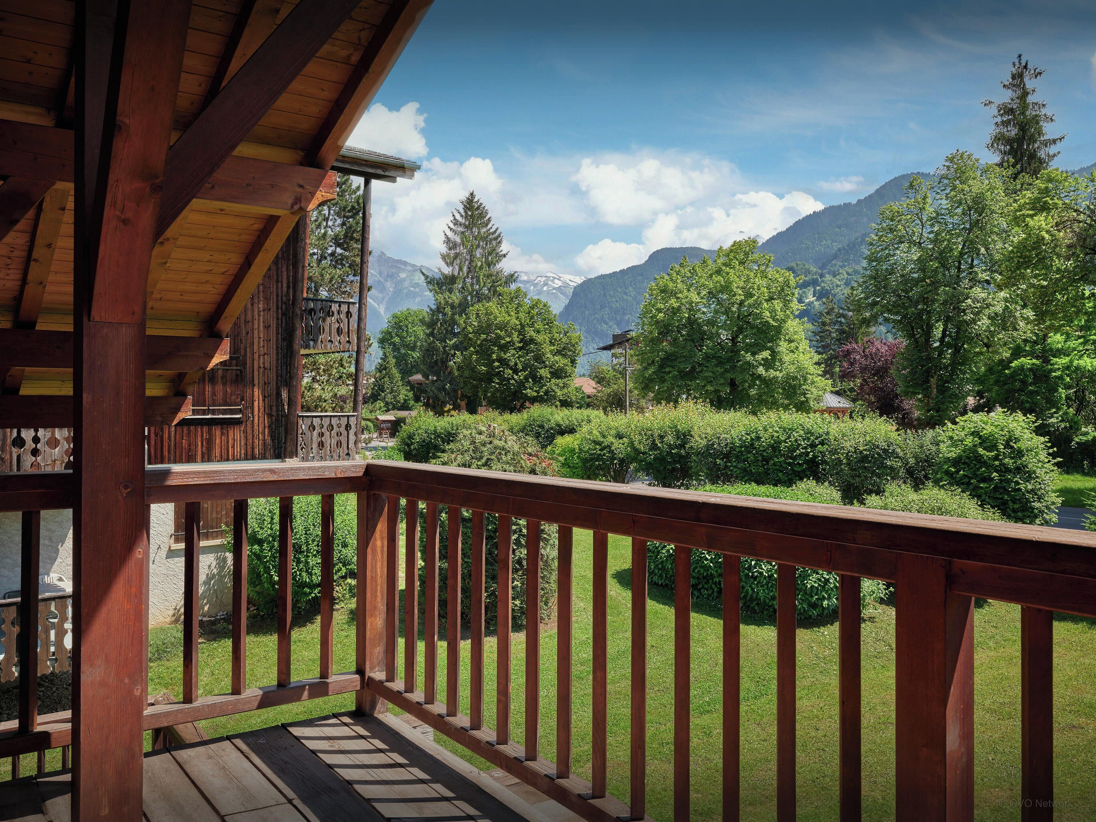 A wooden balcony with a view of a mountainous landscape and a well-maintained garden.
