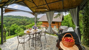A covered outdoor dining area with stone flooring, metal chairs, a table set with plates and a vase of flowers, and a basket of fruit. - Cozy home in Gorkovec (Gorkovec)