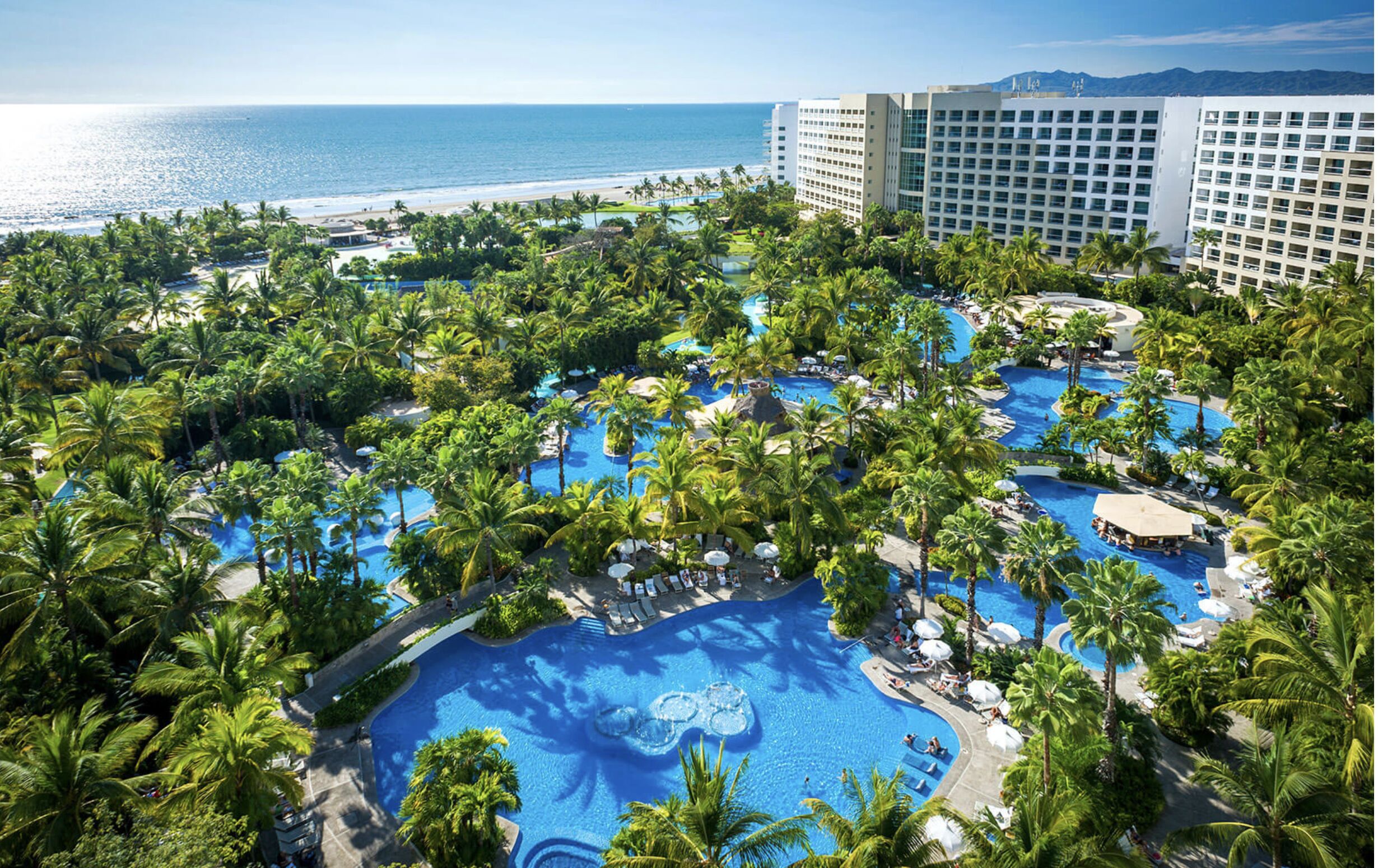 Un resort con varias piscinas rodeado de palmeras, una playa y vistas al océano.