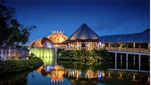 A resort with a thatched-roof building, a modern structure with a glass facade, and a bridge over a reflective water body at night. - Cozy retreat, has something for everyone,property has golf course and lazy river (Las Jarretaderas)