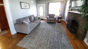 Een woonkamer met een grijze bank, een grijze fauteuil, een open haard en een verwelkomingsbord in een lijst aan de muur.