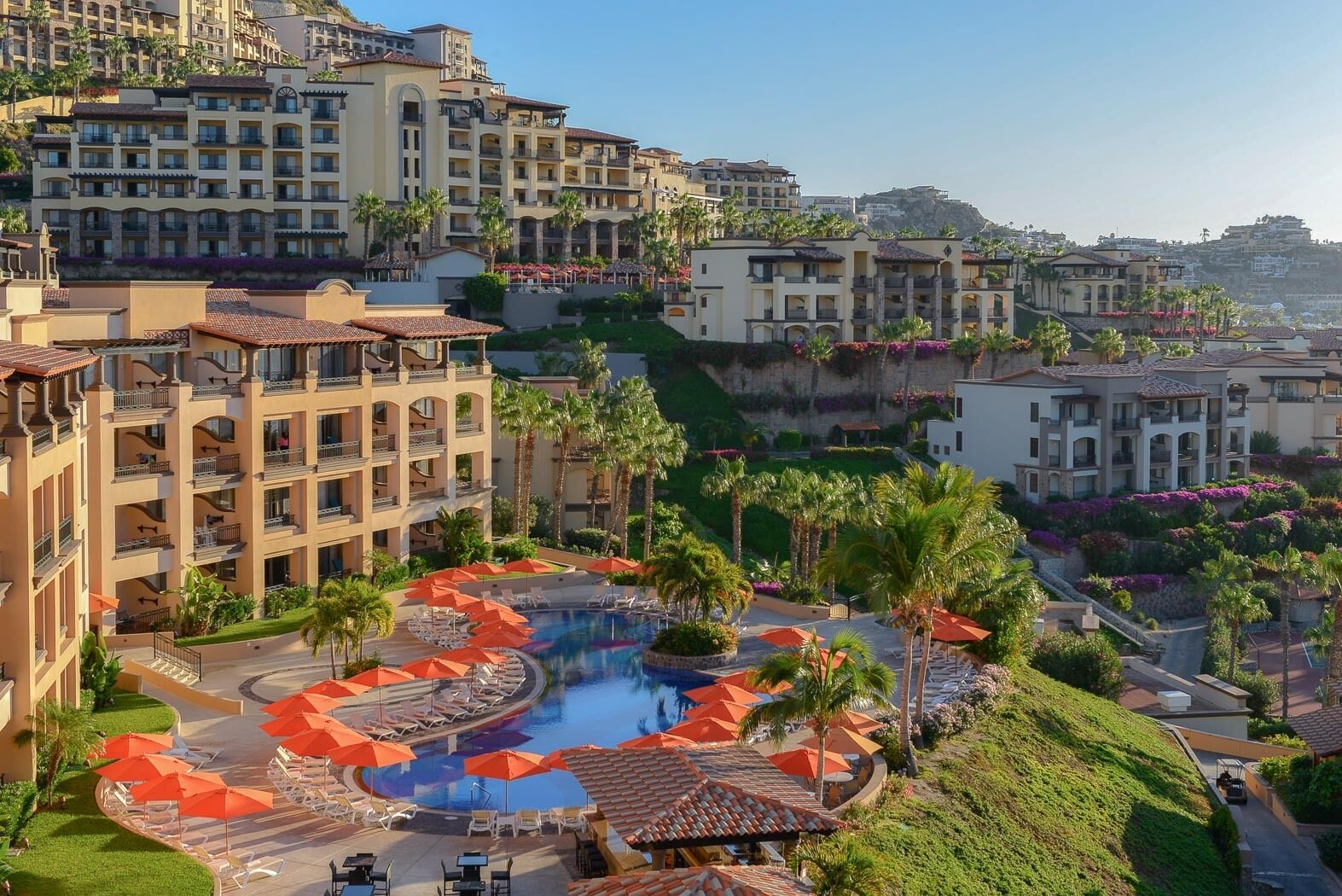 A resort with a central pool surrounded by lounge chairs and umbrellas, multiple hotel buildings, and a hillside backdrop.