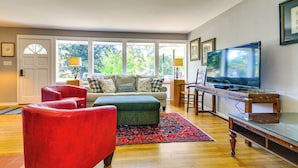 Sala de estar con sofá, reposapiés, sillón rojo, televisor y ventana grande con vistas a la vegetación.
