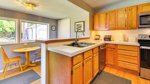 Una cocina con armarios de madera, un lavamanos de acero inoxidable, un microondas y un tostador. Hay una pequeña mesa de comedor con sillas y una ventana que proporciona luz natural.