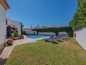 A pool area with lounge chairs, a small garden, and a white building with an archway. - Libertos V4 - VIP Properties by Interhome (Albufeira)
