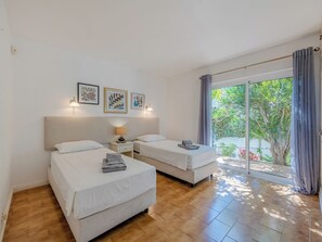 A hotel room with two beds, a small bedside table with a lamp, and a large sliding glass door leading to a balcony with greenery. - Libertos V4 - VIP Properties by Interhome (Albufeira)