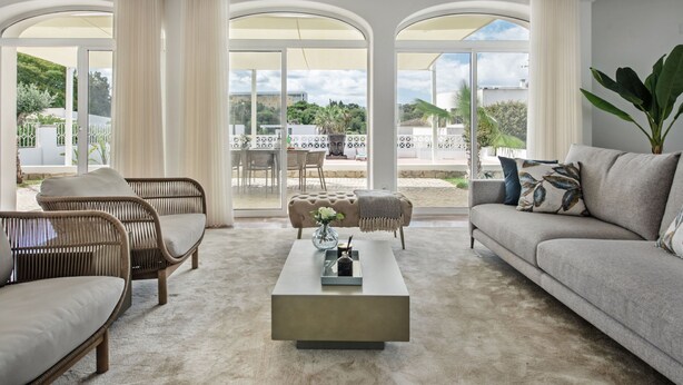 A spacious living room with a large sofa, a coffee table, and wicker chairs. There are large windows providing a view of the outdoors.