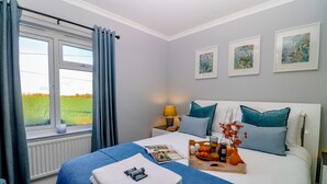 Un lit bien fait, recouvert d’une couverture bleue, une table d’appoint avec des éléments pour le petit-déjeuner et une fenêtre avec des rideaux.