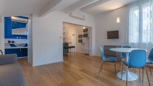 Een moderne woonkamer met een bank, eettafel met stoelen, een televisie en een keukenhoek met een spoelbak en krukjes.