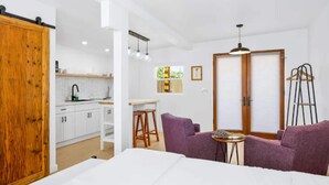 Una habitación moderna con una cama, sofás morados, una mesita y una zona de cocina con armarios blancos y una puerta de madera.
