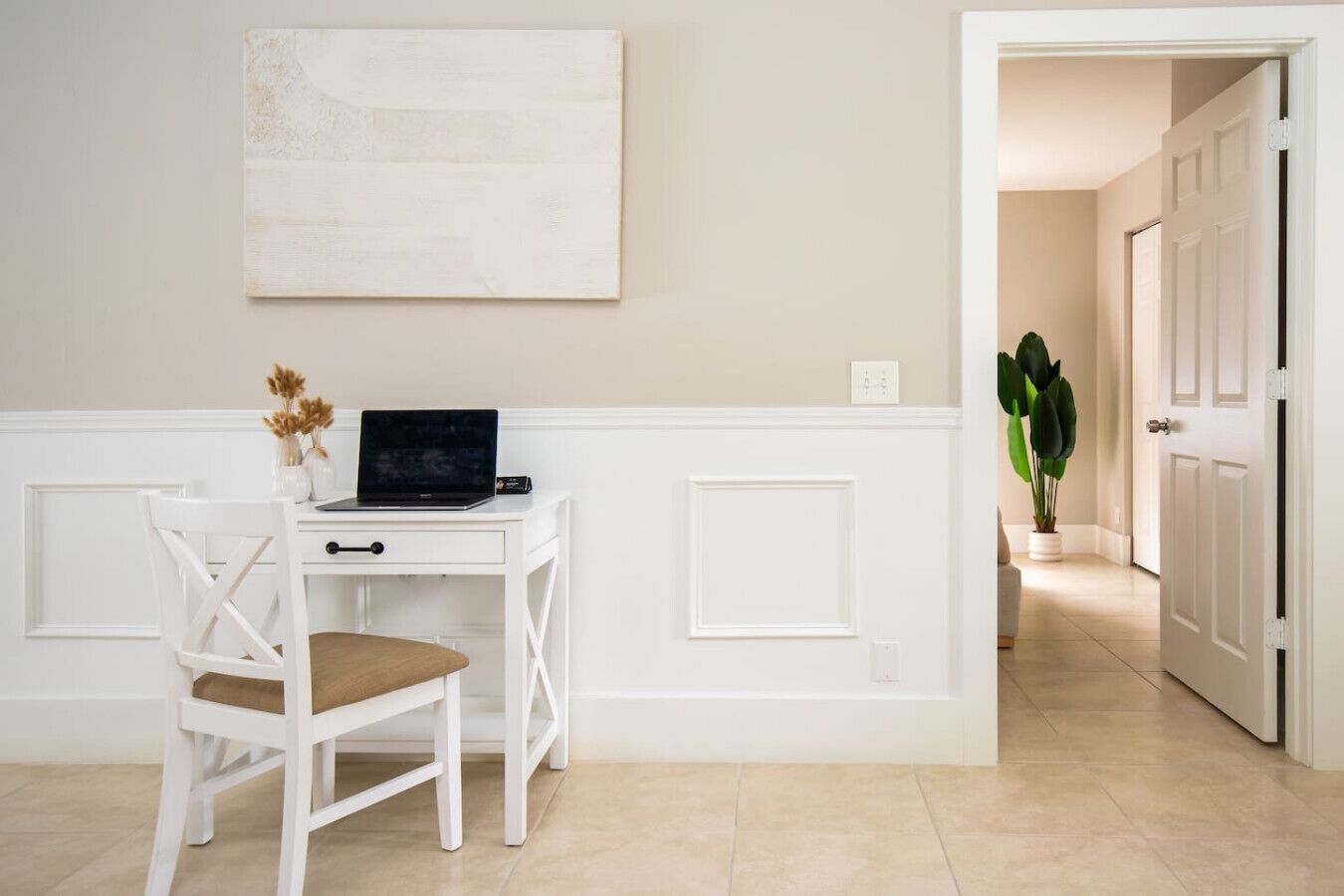 A white desk with a laptop, a chair, and a vase with dried flowers.