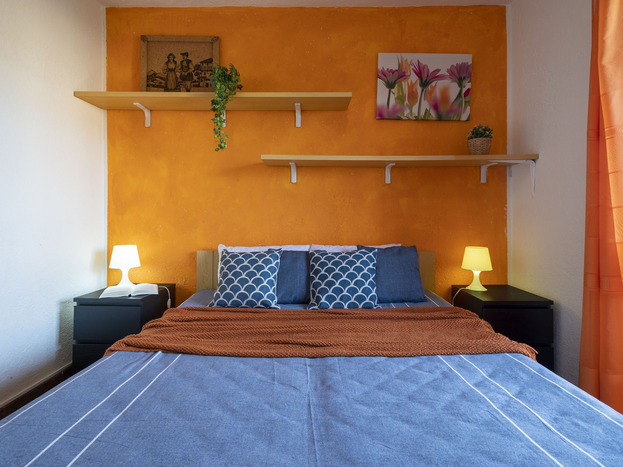 A bedroom with a bed, two bedside tables, two lamps, a blue blanket, a brown throw, a wall-mounted shelf with a plant, and a framed picture.