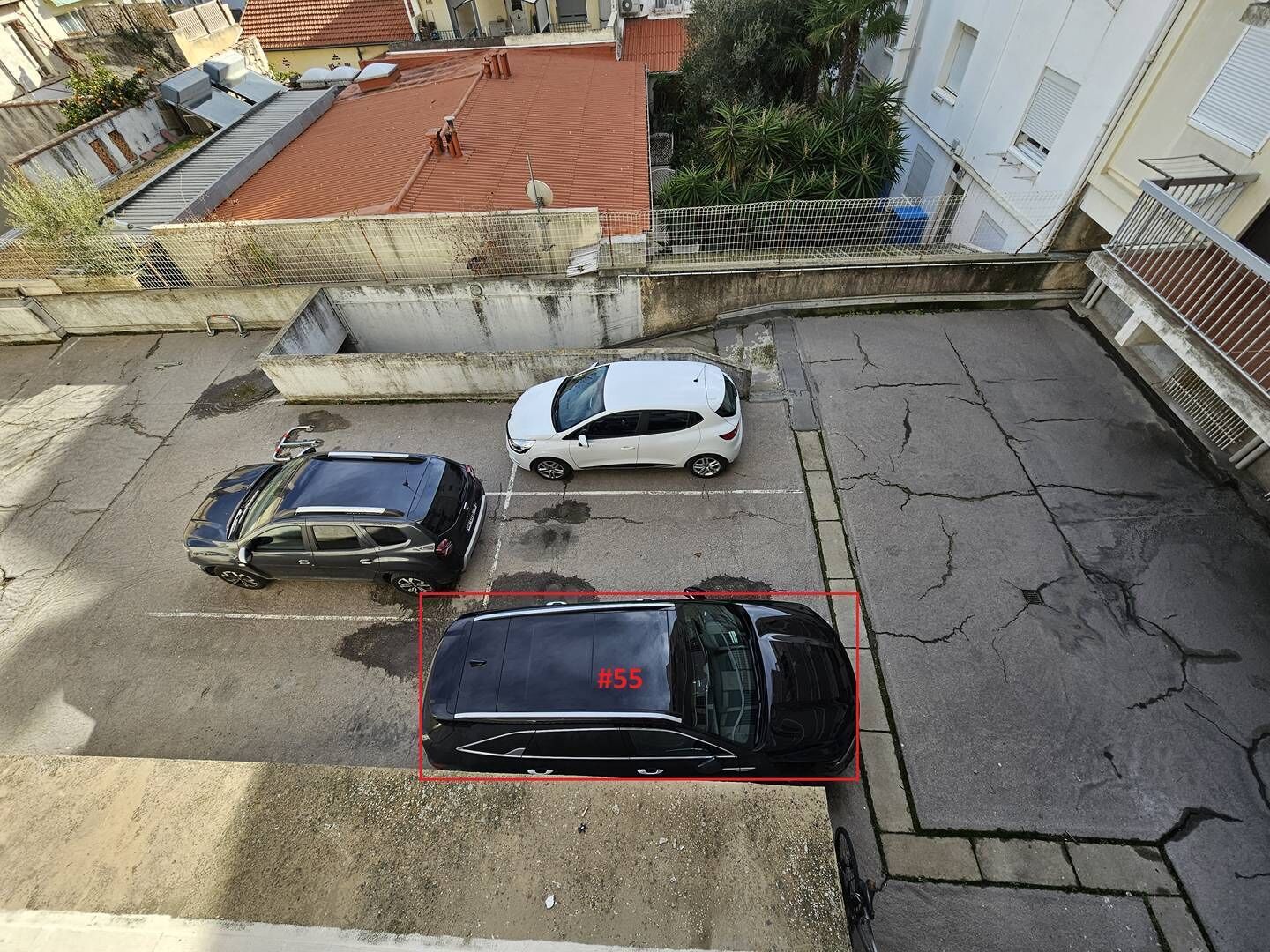 Ein Parkplatz mit drei Autos, einer roten Markierung und einem Gebäude mit rotem Dach im Hintergrund.