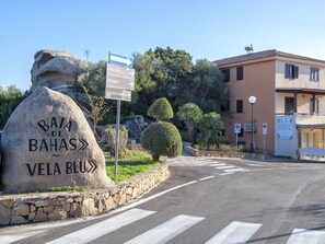 Un panneau en pierre portant les inscriptions « BAIA DE BAHAS » et « VELA BLU », une route menant à un bâtiment avec une enseigne, et un ciel dégagé.