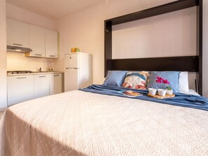A hotel room with a bed, a bedside table with two cups and a tray, a kitchen area with a stove and cabinets, and a black-framed headboard. - D3 - 2 - Baia de Bahas by Interhome (Golfo di Marinella)