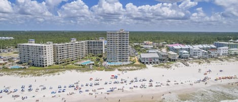 A beachfront hotel with multiple buildings, a swimming pool, and a sandy beach with umbrellas and people.