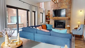A modern living room with a stone fireplace, a large sofa, a television, and a balcony with a wooden railing. - Close to Lifts | Shuttle Stop | Renovated | Windswept 5 (Angel Fire)