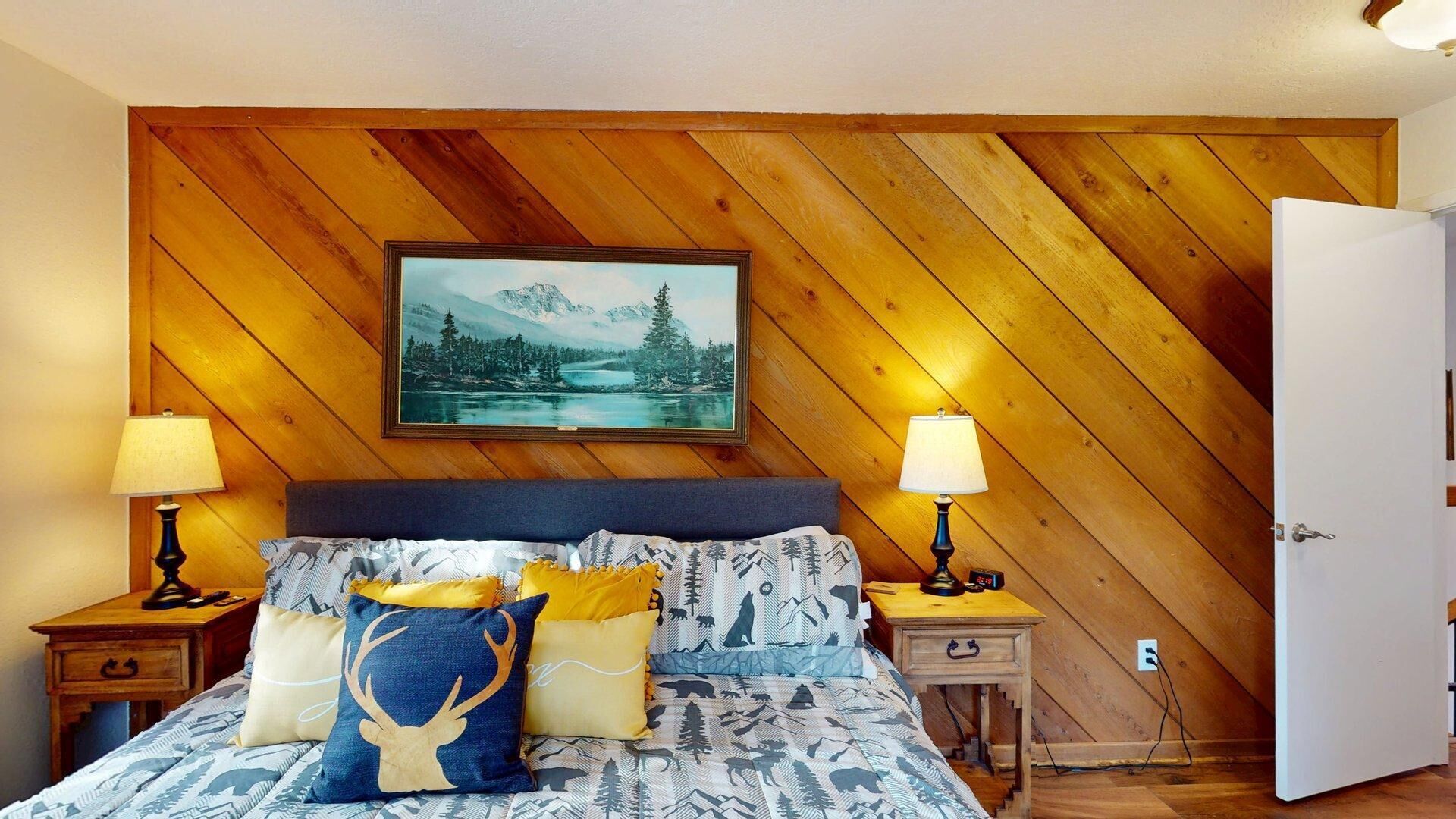 A bedroom with a wooden sloped ceiling, a bed with patterned bedding, two bedside tables with lamps, and a painting above the bed.