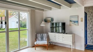 A room with a large sliding glass door leading to a garden, a white cabinet with drawers, a television on top, and a chair with a patterned cover. - 4 bedroom cozy home in Beddingestrand (Beddingestrand)