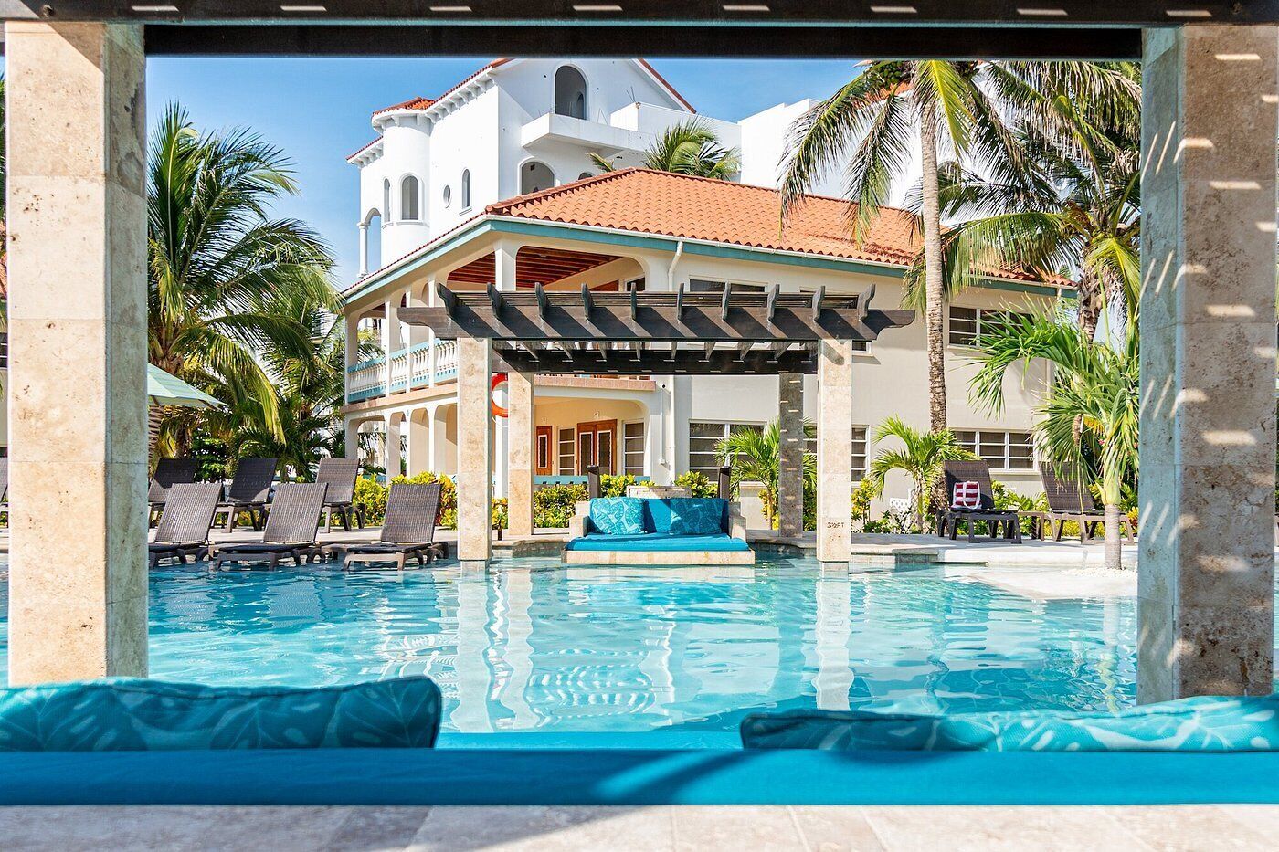 Zona de piscina con sillones de descanso y un espacio de estar cubierto, rodeada de palmeras y un edificio con techo de tejas rojas.